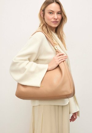 Pitkähiuksinen vaaleatukkainen nainen, joka pukeutunut kermaiseen neulepaita ja hameeseen, ja kantaa suurta ruskeaa nahkaista olkalaukkua, jossa on kullanvärisiä yksityiskohtia.