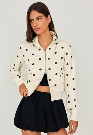 Kvinde iført en hvid lynlåsjakke med sorte polkaprikker og sorte løse shorts, stående mod en ensfarvet lys baggrund.