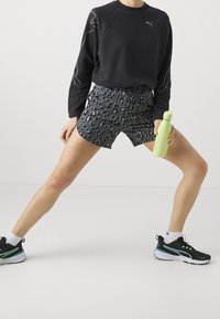 Felpa nera con il logo Puma, pantaloni corti neri a righe e scarpe da ginnastica nere con dettagli verdi. Tiene una borraccia verde chiaro.
