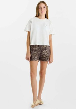 Jonge vrouw staand in wit cropped T-shirt, korte broek met luipaardprint en gouden platte schoenen tegen een effen witte achtergrond.