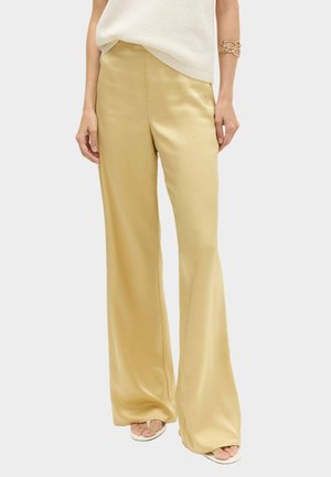 Frau trägt beige Satin-Hosen mit weitem Bein, weißes ärmelloses Oberteil, weiße Sandalen mit offenen Zehen und ein goldenes Gliederarmband am linken Arm.