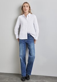 Camicia bianca a maniche lunghe con design a bottoni, abbinata a jeans blu a gamba dritta e scarpe nere. Tessuto liscio e vestibilità casual.
