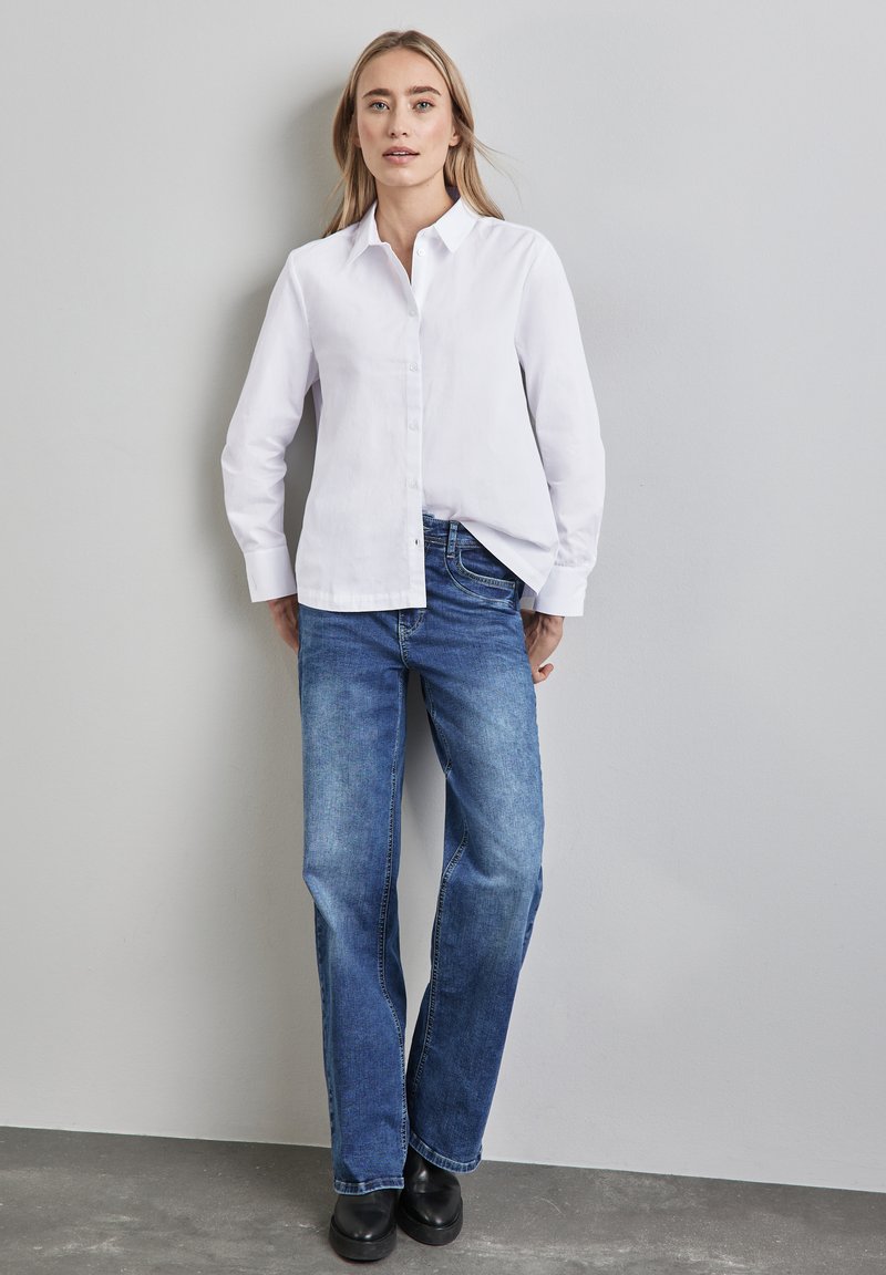 Camicia bianca a maniche lunghe con design a bottoni, abbinata a jeans blu a gamba dritta e scarpe nere. Tessuto liscio e vestibilità casual.