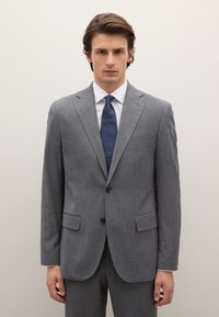 Grau maßgeschneiderter Anzug mit Kerbkragen, zwei Vorder Taschen und dunkelblauer Krawatte mit weißen Punkten, getragen über einem weißen Hemd. Glatte Textur.