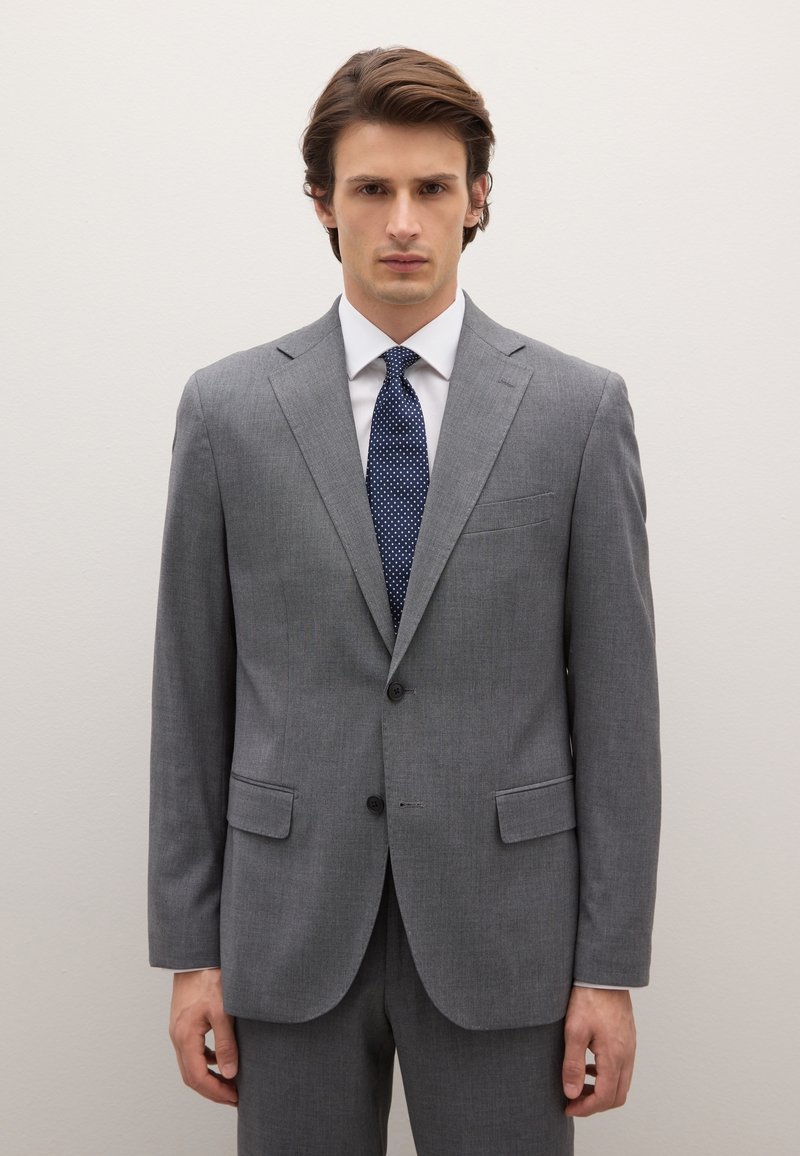 Grau maßgeschneiderter Anzug mit Kerbkragen, zwei Vorder Taschen und dunkelblauer Krawatte mit weißen Punkten, getragen über einem weißen Hemd. Glatte Textur.