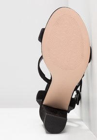 Sandale en daim noire à talon bloc bas, semelle beige avec motif ondulé et bride de cheville avec fermeture à boucle.