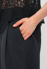 Main reposant dans la poche d'un pantalon noir porté avec un haut en dentelle noire, montrant le contraste de texture entre les tissus.