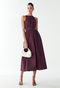 Burgundska halter obleka z nabranim pasom in silhueto v obliki črke A, v kombinaciji s kremno torbico z okruglim ročajem in zlatimi detajli.