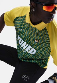 La chemise polo Nike présente un col et des manches jaunes, un corps à motifs verts avec des accents jaunes. Elle est assortie à un pantalon noir arborant un logo.