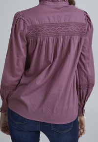 Maglietta lunga viola con scollo arricciato, dettagli in pizzo sulla schiena e design plissettato, realizzata in un tessuto leggero.