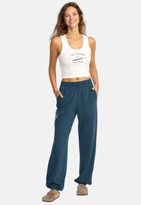 Vit, åtsittande linne med blå text; lösa, mörk tealfärgade sweatpants med sidofickor och logodetalj. Avslappnad, bekväm outfit.