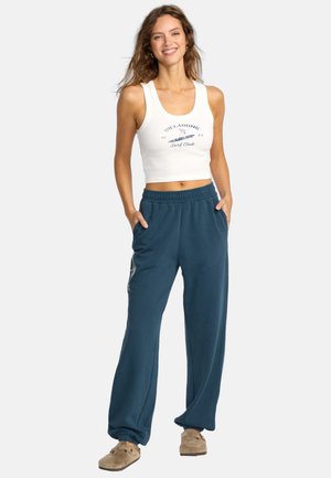 Witte, aansluitende tanktop met blauwe tekst; losse, donkerblauwe sweatpants met zijzakken en logodetail. Casual, comfortabele outfit.