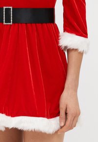 Rdeča žametna obleka z belimi umetnimi krznom obrezanimi rokavi in robom, ki vključuje širok črn pas z zaponko iz kristalov ter prikazuje prilegajoč dizajn.