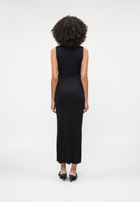 Μαύρο μίνι φόρεμα maxi χωρίς μανίκια, εφαρμοστό, με λεία υφή, που διαθέτει ψηλό λαιμό και ίσια κουπ, φορεμένο με μαύρα τακούνια.