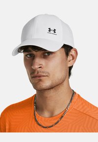 Casquette blanche avec une finition texturée, visière courbée et un logo Under Armour noir à l'avant. Portée avec une chemise à motif orange et une chaîne en argent.