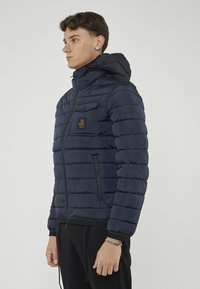 Giacca a soffietto blu navy con cappuccio, design trapuntato, tasche frontali con zip e una toppa sul petto. Realizzata in materiale leggero e lucido.