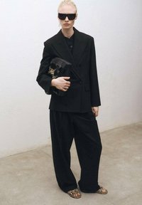 Černé oversized sako s odpovídajícími širokými kalhotami, doplněné černou kabelkou a tmavými slunečními brýlemi. Ozdobené vzorovanými sandály.