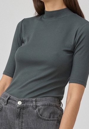 Rippiertes, kurzärmeliges T-Shirt in Dunkelgrün mit hohem Kragen. Kombiniert mit hoch taillierten, grauen Jeans aus Denim mit einer Vordertasche.