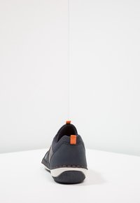 En ensam mörkblå sneaker med orange dragflik och sömmar, visad bakifrån på en vit yta mot en vit vägg.