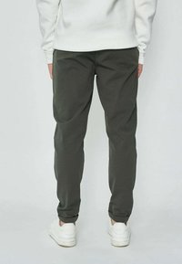 Personne portant un pantalon décontracté vert foncé et des baskets blanches, se tenant dos à la caméra sur un fond clair uni.