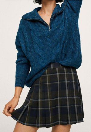 Femme portant un pull bleu en maille torsadée à fermeture éclair quart de zip et une jupe plissée à carreaux foncés, tenant l'ourlet de la jupe d'une main.