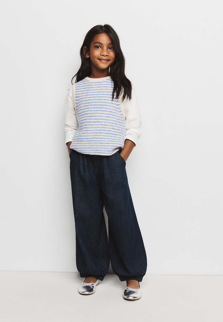 Fille aux longs cheveux foncés portant une chemise blanche à manches longues, un gilet rayé bleu et blanc, un pantalon ample foncé et des chaussures argentées, debout les mains dans les poches.