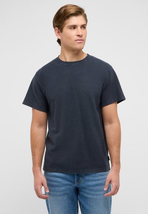 Young man in a plain dark gray T-shirt and blue jeans standing against a light gray background, looking to the side.