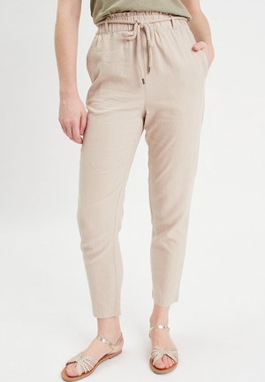Pantalon beige taille haute à coupe fuselée avec taille à cordon, porté avec des sandales plates à sangle tissée métallique. Une main dans la poche, debout sur fond blanc.