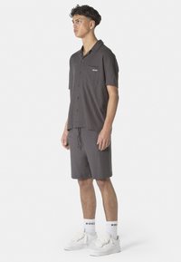 Graues kurzärmeliges Hemd mit Knöpfen und einer Brusttasche, kombiniert mit passenden Shorts. Das Modell trägt weiße Sneakers und Socken mit Logo.