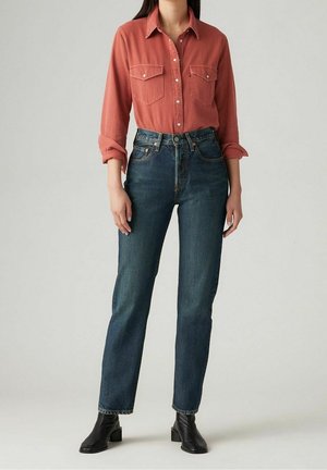 Femme portant une chemise rouille boutonnée, un jean taille haute bleu foncé et des bottines noires, debout devant un fond uni.