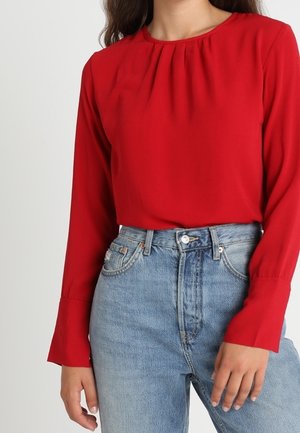 Red long-sleeve blouse with gathered neckline, smooth texture, paired with high-waisted light blue jeans featuring a faded wash and classic button closure.