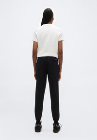 Persoană văzută din spate, purtând un tricou alb cu mâneci scurte, pantaloni negri de trening și adidași negri, pe un fundal simplu, deschis.