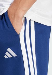 Blauwe sportbroeken met witte zijstrepen en een Adidas-logo. De stof lijkt zacht en iets rekbaar, met een comfortabele pasvorm.