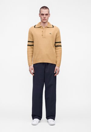 Hombre con un suéter de canalé color beige con rayas negras en las mangas, pantalones anchos de pierna oscura y zapatillas blancas, de pie frente a un fondo blanco.