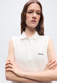 Νεαρή γυναίκα με μακριά καστανά μαλλιά, φορούσα λευκή αμάνικη πόλο μπλούζα με το λογότυπο "Barbour", σταυρωμένα χέρια, ουδέτερη έκφραση, απλό ανοιχτόχρωμο φόντο.