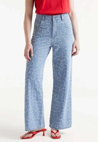 Vaqueros de talle alto azul claro con bordado floral, diseño de pierna ancha y detalles deshilachados. Combinados con sandalias de tacón rojas.