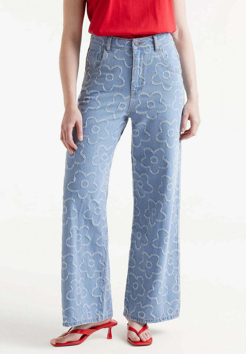 Vaqueros de talle alto azul claro con bordado floral, diseño de pierna ancha y detalles deshilachados. Combinados con sandalias de tacón rojas.