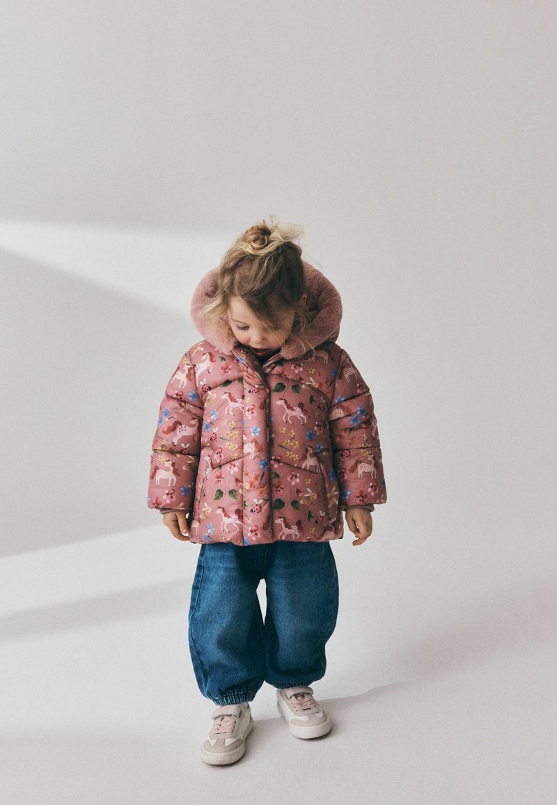 Jeune enfant portant une doudoune rose à motif floral et licorne, un jean bleu et des baskets blanches, regardant vers le bas sur un fond uni.
