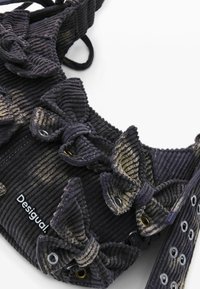 Borsa in velluto a coste nera con dettagli arricciati e accessori dorati. Texture e design distintivi con un'etichetta brandizzata. Dettagli includono occhielli lungo la tracolla.