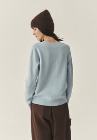 Heleheline kootud sviiter ribide ääristega, kantud pruunide pükste ja tumeda pruuni mütsiga. Keskendu pehmele tekstuurile ja vabale lõikele.