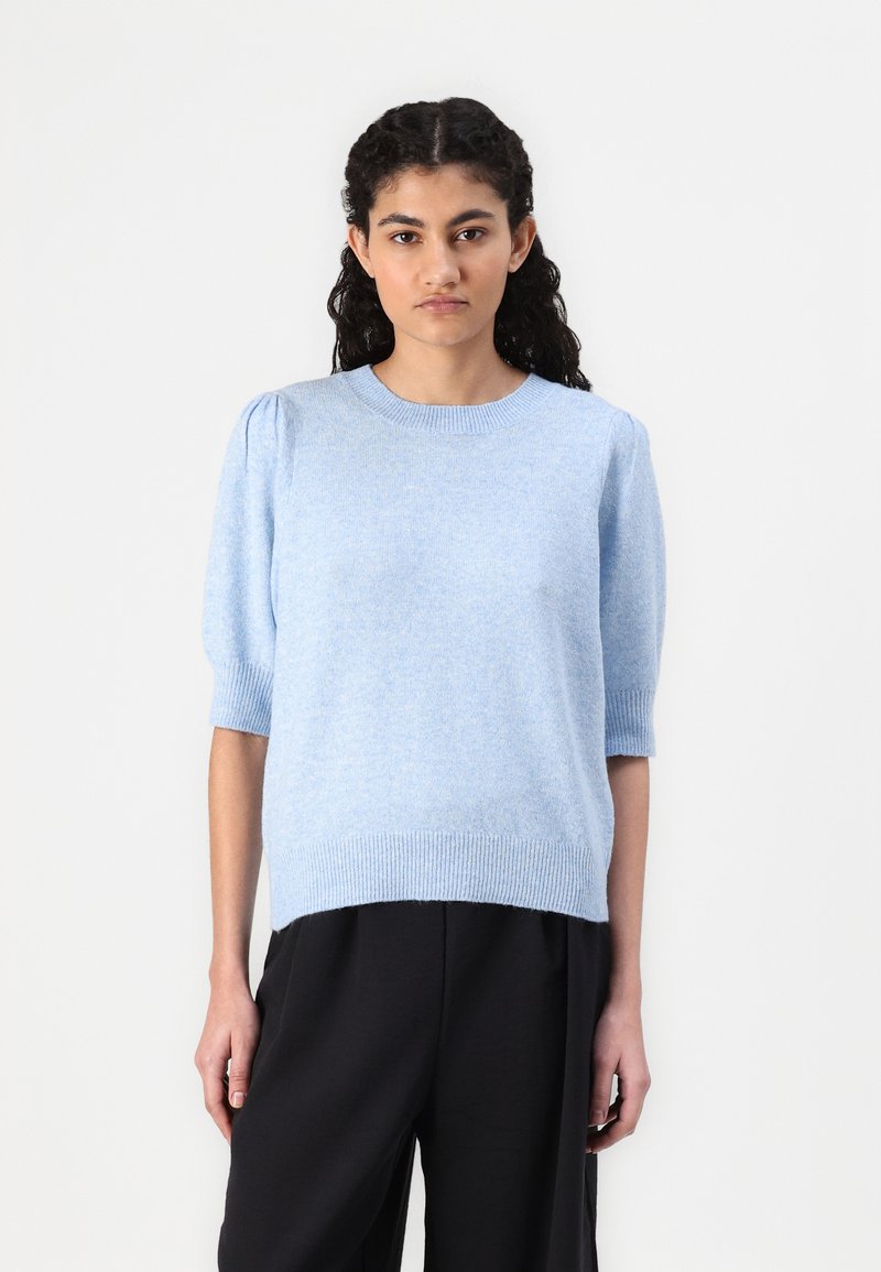 Suéter tricotado azul claro com mangas curtas bufantes, gola e bainha caneladas, corte solto, usado sobre calças pretas. Textura suave.