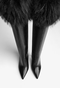 Čierne kožené čižmy siahajúce po kolená s ostrými špičkami, s nadýchaným čiernym kožušinovým lemom na vrchu a elegantným bočným zipsom.