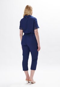 Navy blauwe jumpsuit met korte mouwen, elastische tailleband en cropped broeken. Heeft kantafwerking op de mouwen en een knoopdetail op de rug.