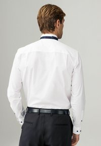 Camisa blanca de manga larga con cuello en contraste en navy. Botones de puño negros visibles. Llevada con pantalones oscuros y un cinturón negro.