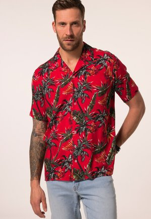 Mann mit Bart trägt ein rotes kurzärmeliges Hemd mit tropischem Blatt- und Palmenmuster und hellblaue Jeans vor schlichtem Hintergrund.