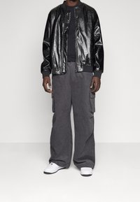 Černá lesklá bomber bunda s krokodýlím vzorem přes přiléhavé černé tričko, doplněná volnými šedými cargo kalhotami a bílými teniskami.