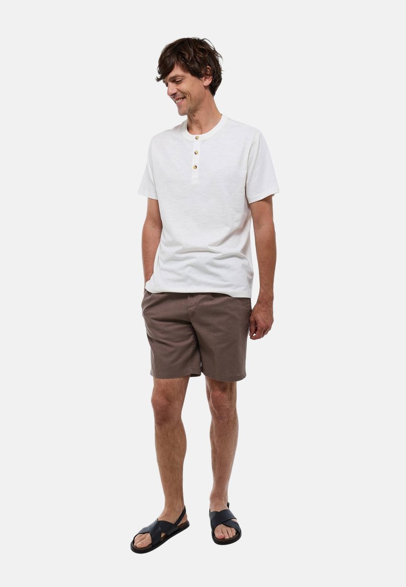 Chemise henley blanche à manches courtes avec trois boutons en bois, short marron et sandales noires. Coupe décontractée avec une texture douce.