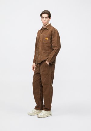 Braune Cordjacke mit Knopfverschluss und einer Brusttasche, kombiniert mit passenden Hosen. Die Schuhe sind hellfarbene, strukturierte Sneakers.