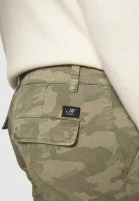 Camouflage cargo-byxor i olivgrön med mjuk textur. Har en bakficka med en svart etikett som säger "MASON'S." Tillverkat av bomull.