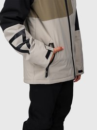 Persona che indossa una giacca beige, nera e oliva con una mano nella tasca laterale con zip e pantaloni neri su uno sfondo neutro.